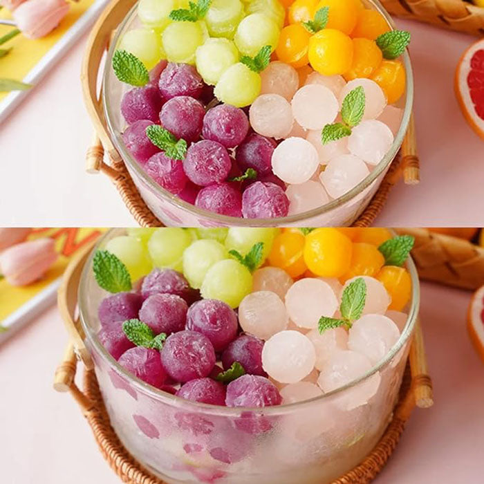 The image shows a clear glass bowl filled with colorful round ice cubes and fruit-shaped ice balls. The ice cubes are arranged in two layers, with vibrant colors including purple, green, yellow, and pale shades of pink and white. Fresh mint leaves are placed among the ice, adding a touch of greenery. The bottom half of the bowl displays a similar arrangement of ice cubes with slightly varied positioning. The overall presentation appears fresh and inviting, suggesting a refreshing treat.
