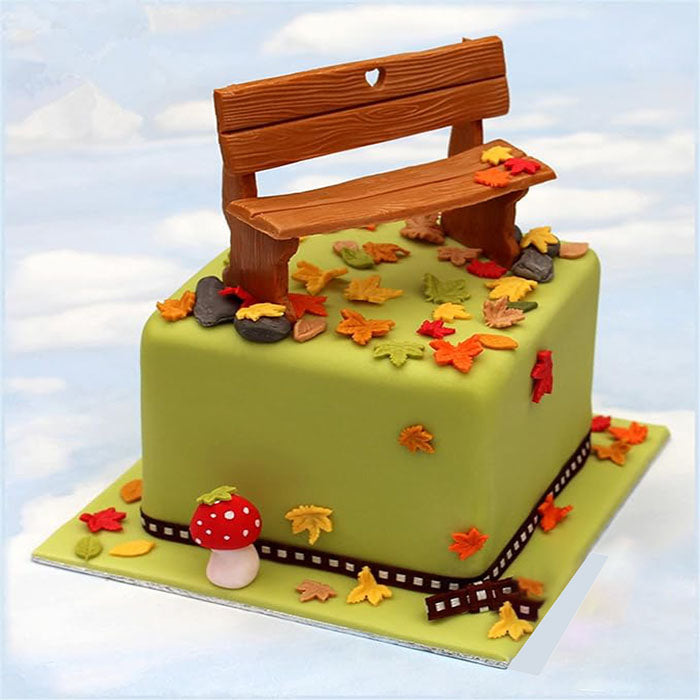A decorative cake shaped like a green cube, topped with colorful fondant maple leaves in red, orange, and yellow. A small brown bench made of fondant sits on top, with a heart-shaped cutout. There are rocks and a red and white spotted mushroom near the base of the cake, which is bordered with a film strip design. The background features a blue sky.
