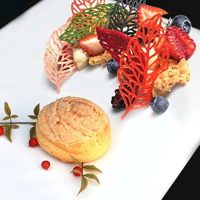 This image features a beautifully arranged dessert on a white plate. Prominent in the display are several decorative hollow leaf-shaped elements in various colors, including red, green, and orange. Alongside the leaves, there are fresh fruits such as strawberries and blueberries, as well as smaller decorative items resembling red berries and leafy sprigs. In the center of the plate sits a small, round, golden-brown pastry, adding to the overall colorful and artistic presentation of the dish.