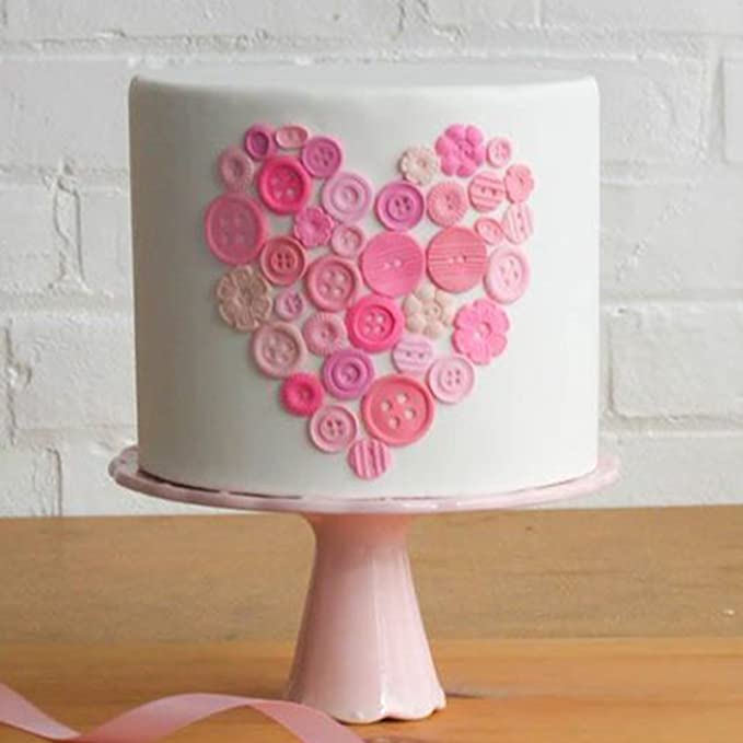 A decorated cake featuring a smooth, white frosting surface with a heart shape made of colorful faux buttons arranged in shades of pink and light pink. The cake is displayed on a pink pedestal stand with a ribbon draped across the table beneath it. The background consists of a neutral brick wall.