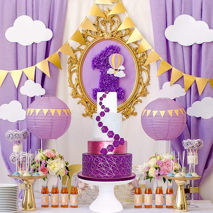 An elegantly decorated birthday cake display featuring a three-tiered cake. The cake has a white top tier, a middle tier with a purple ombre effect, and a bottom tier with a geometric pattern in dark purple. The cake is adorned with hexagon shapes in varying shades of purple. The background is decorated with purple curtains, golden bunting, and paper lanterns. There are white clouds and a framed number one made of purple decorations, along with floral arrangements and colorful treats on the table.