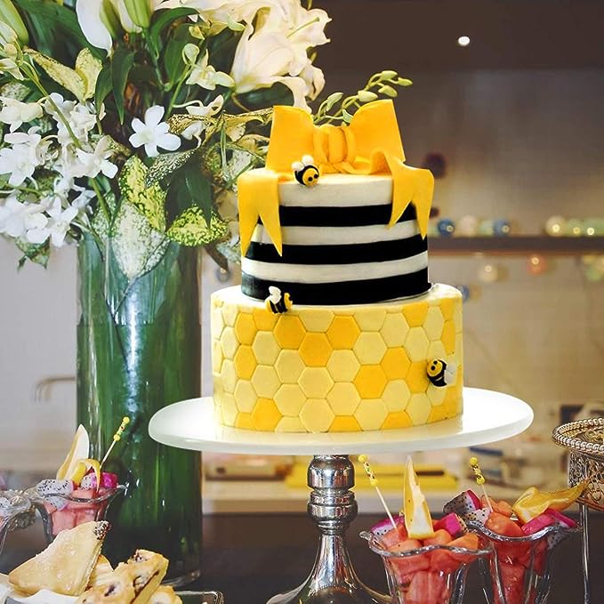 A two-tiered cake is displayed on a cake stand. The top tier is black and white striped, adorned with a yellow bow and small bee decorations. The bottom tier has a honeycomb pattern in shades of yellow, also with small bee details. Surrounding the cake are various colorful desserts in glass cups and a tall vase filled with white flowers in the background.