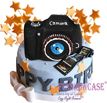 A decorative cake designed to resemble a black camera, featuring a round lens with a blue center. The cake is placed on a light blue base with "HAPPY BIRTHDAY" written in bold letters. Surrounding the cake are golden star-shaped decorations, adding a festive touch. The scene includes a few small, edible photo prints placed on the cake, enhancing the camera theme.