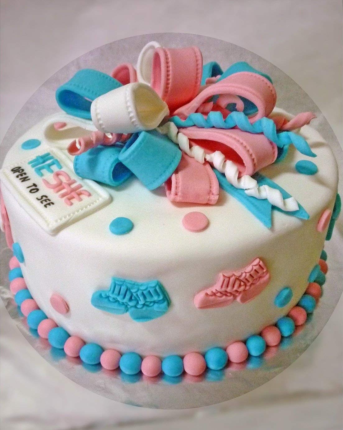 A round cake covered in smooth white fondant features a colorful decorative design. The top is adorned with a bow made of blue, pink, and white ribbons. The cake has small circular decorations in pastel pink and blue. There are also small boot shapes in blue and pink on the sides of the cake. A label on the cake reads "HE/SHE OPEN TO SEE." The bottom of the cake is encircled with a row of alternating pink and blue pearl-like decorations.