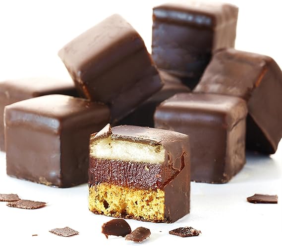 A close-up image of square-shaped chocolate confections is displayed. Several dark chocolate-covered cubes are stacked in the background, while one cube is cut in half to reveal its layered interior. The exposed section shows three distinct layers: a chocolate top layer, a fruit or jelly middle layer, and a crumbly base layer, all resting on a clean white surface. Small chocolate shavings are scattered around.