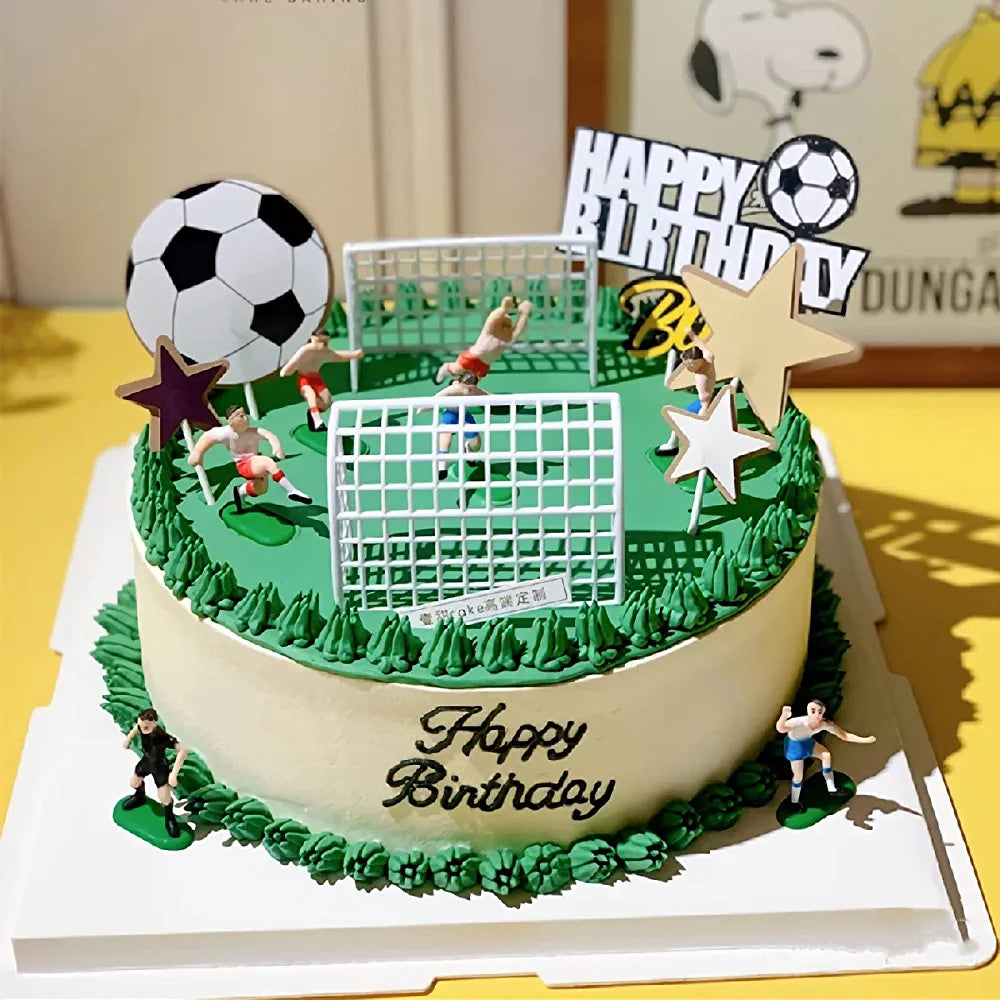 A round birthday cake themed for soccer, decorated with green icing and a grass-like texture. On top, there are small figurines representing soccer players in action, two mini goals, and a large black and white soccer ball. The cake also features stars and a banner that reads "Happy Birthday." The cake sits on a white platform against a yellow background.