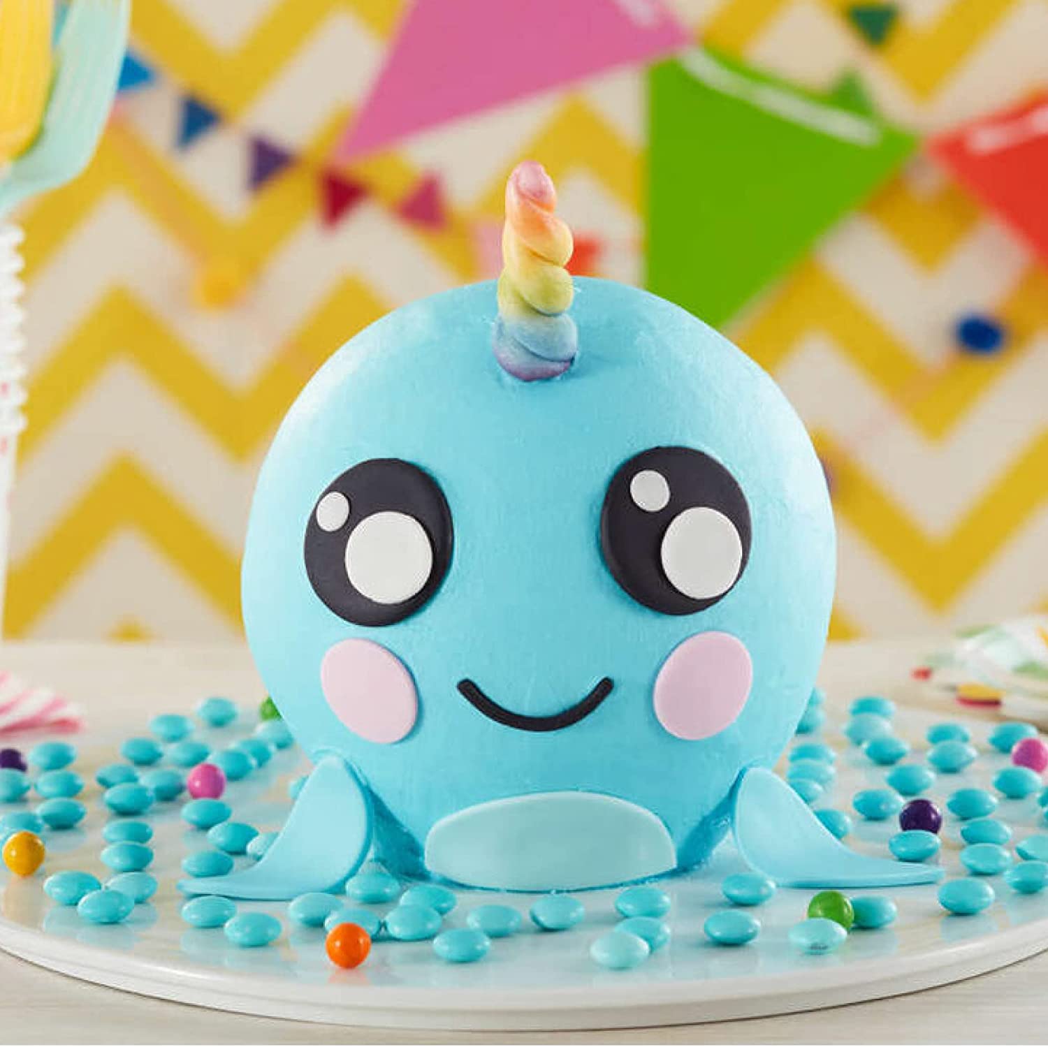 A spherical, bright blue cake designed to resemble a cheerful whale, featuring large black eyes with white highlights and pink cheeks. It has a whimsical rainbow-colored horn on top and small fins at the sides. The cake sits on a white round platter, surrounded by pastel-colored candy pieces, and is set against a colorful, zigzag-patterned background with decorative paper banners in various colors.