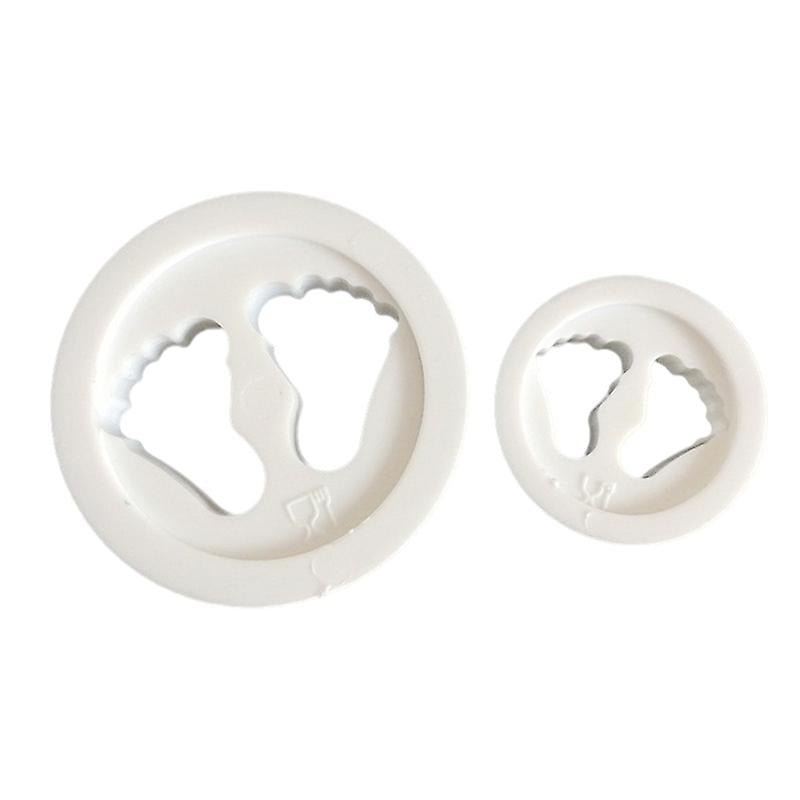 Two cookie cutters shaped like baby feet, one larger and one smaller, both made of white plastic. The larger cutter features the outline of a pair of baby feet, while the smaller one has a similar design but on a smaller scale.