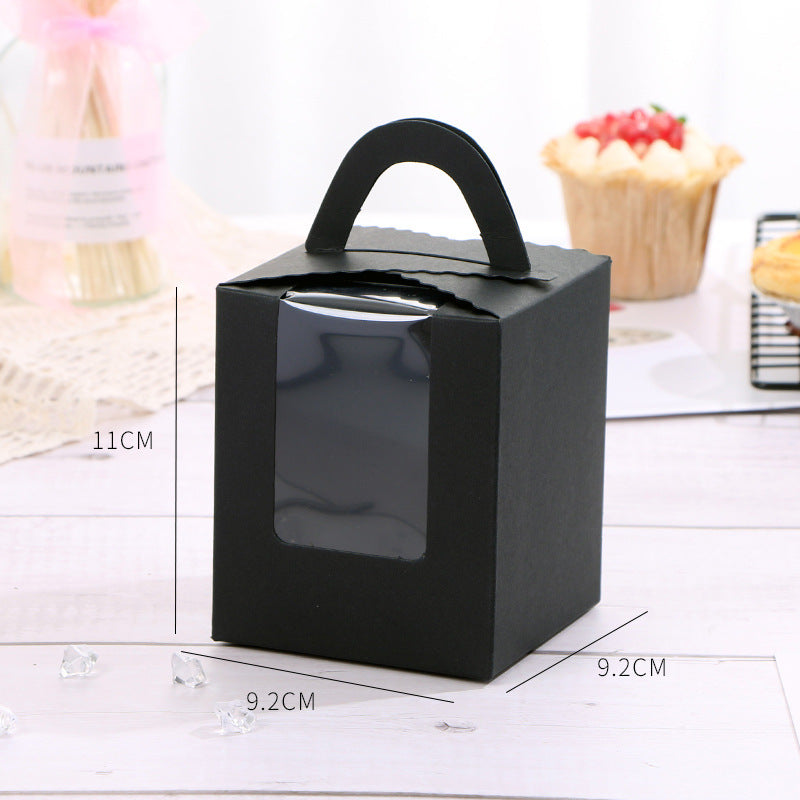 A black cardboard cupcake box with a handle, designed to hold a single portion. The box measures 11 cm in height and 9.2 cm in width. It features a clear window on the front, allowing visibility of the cupcake inside. In the background, there is a decorative cupcake topped with red fruit and a plate of cookies. The setting includes a light-colored wooden surface and some decorative crystals scattered around.