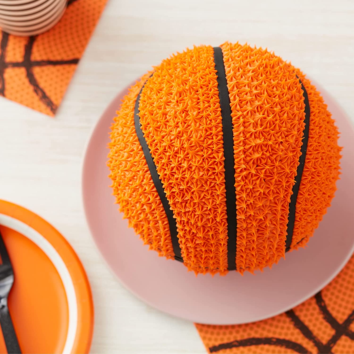 An orange 3D cake shaped like a basketball sits on a pink plate. The cake is detailed with a textured surface resembling basketball dimples and features black icing stripes that mimic the design of an actual basketball. Surrounding the cake are orange-themed party supplies, including a decorative napkin and a colorful plate. The background is a light-colored surface, adding to the festive presentation.