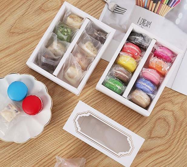 A wooden surface features a white box with a clear top containing ten colorful macarons in various flavors, arranged in two rows. Next to it, a second box holds smaller, individually wrapped confections in different colors and flavors. There is also a decorative white plate with three additional macaron-style treats in red and blue. A printed paper and a fork are visible in the background.