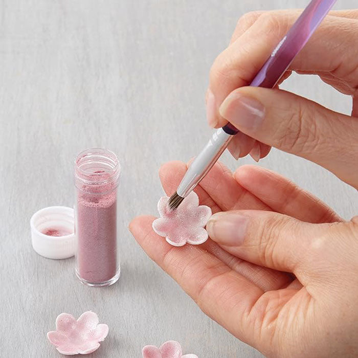 A close-up image shows a person using a decorating brush to apply pink powder to a small flower-shaped decorative piece. A container of pink powder is visible alongside another small lid. The background is a light-colored surface.
