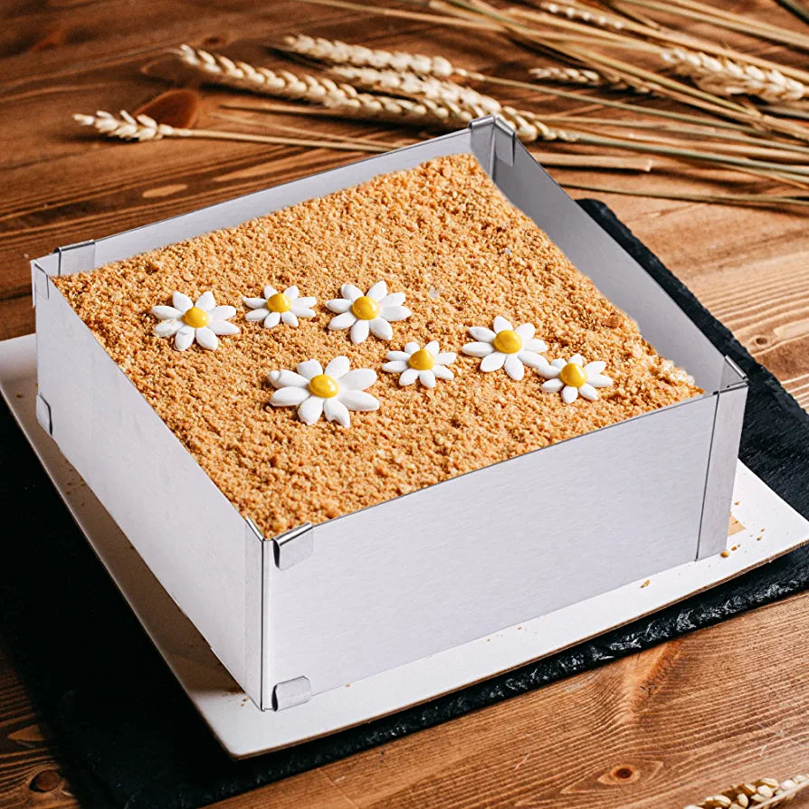 An adjustable square cake ring is placed on a wooden table, featuring a decorated cake inside. The cake has a crumbly topping and is adorned with white flower-shaped decorations, each with a yellow center, arranged in a row. The cake ring itself is made of silver-colored material and fits snugly around the cake, with visible corner connections. The cake is presented on a square white plate that contrasts with the dark surface beneath it, while the background includes decorative wheat stalks.