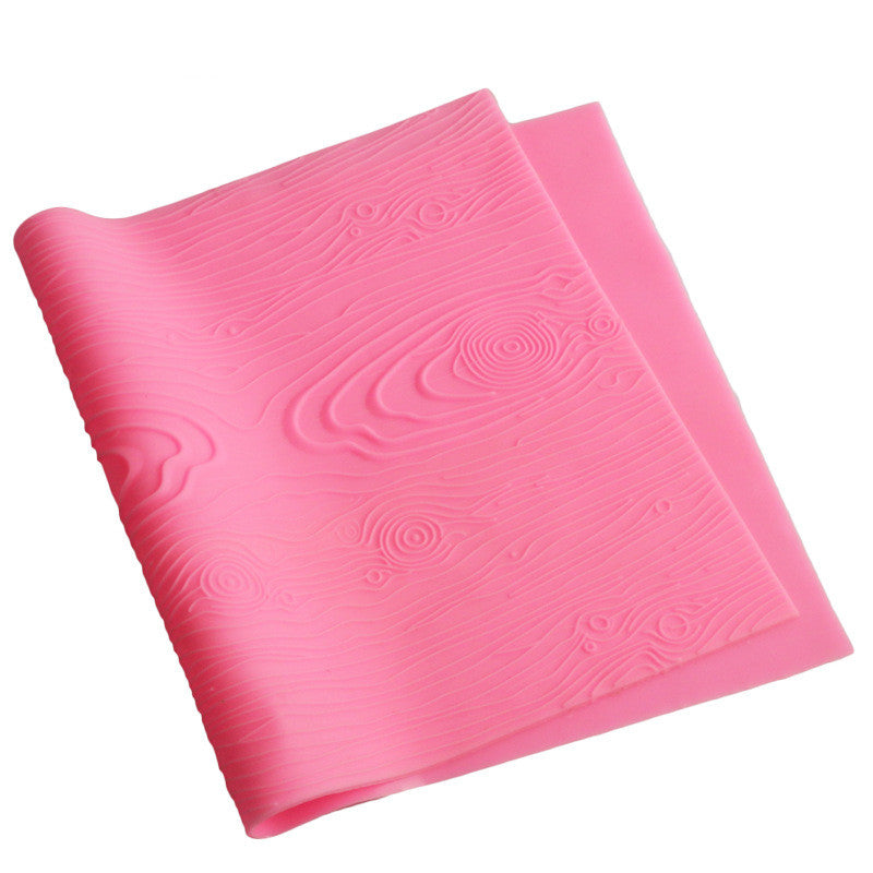 A pink silicone mat designed for creating woodgrain impressions in fondant. The mat features a textured surface that mimics the appearance of wood, with wavy lines and circular patterns to resemble tree rings and wood grain textures. The mat is slightly flexible and can be laid flat for use.