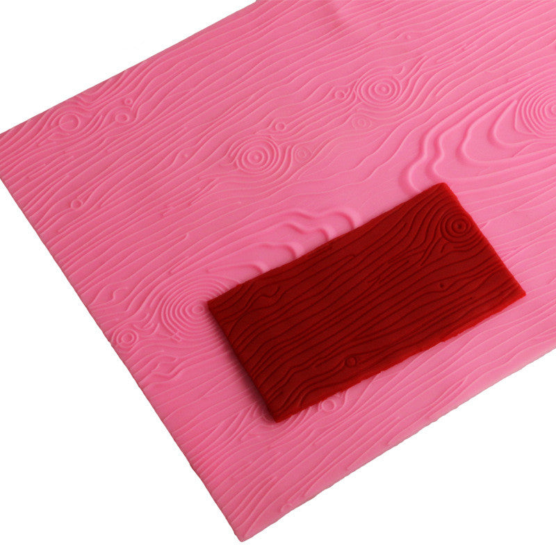 An impression silicone mat is displayed, featuring a textured surface designed to resemble wood grain. The mat is predominantly pink and showcases various wood-like patterns with grooves and knots. A small rectangular section of red silicone, also with wood grain texture, is placed on top of the pink mat. The overall layout emphasizes the mat&