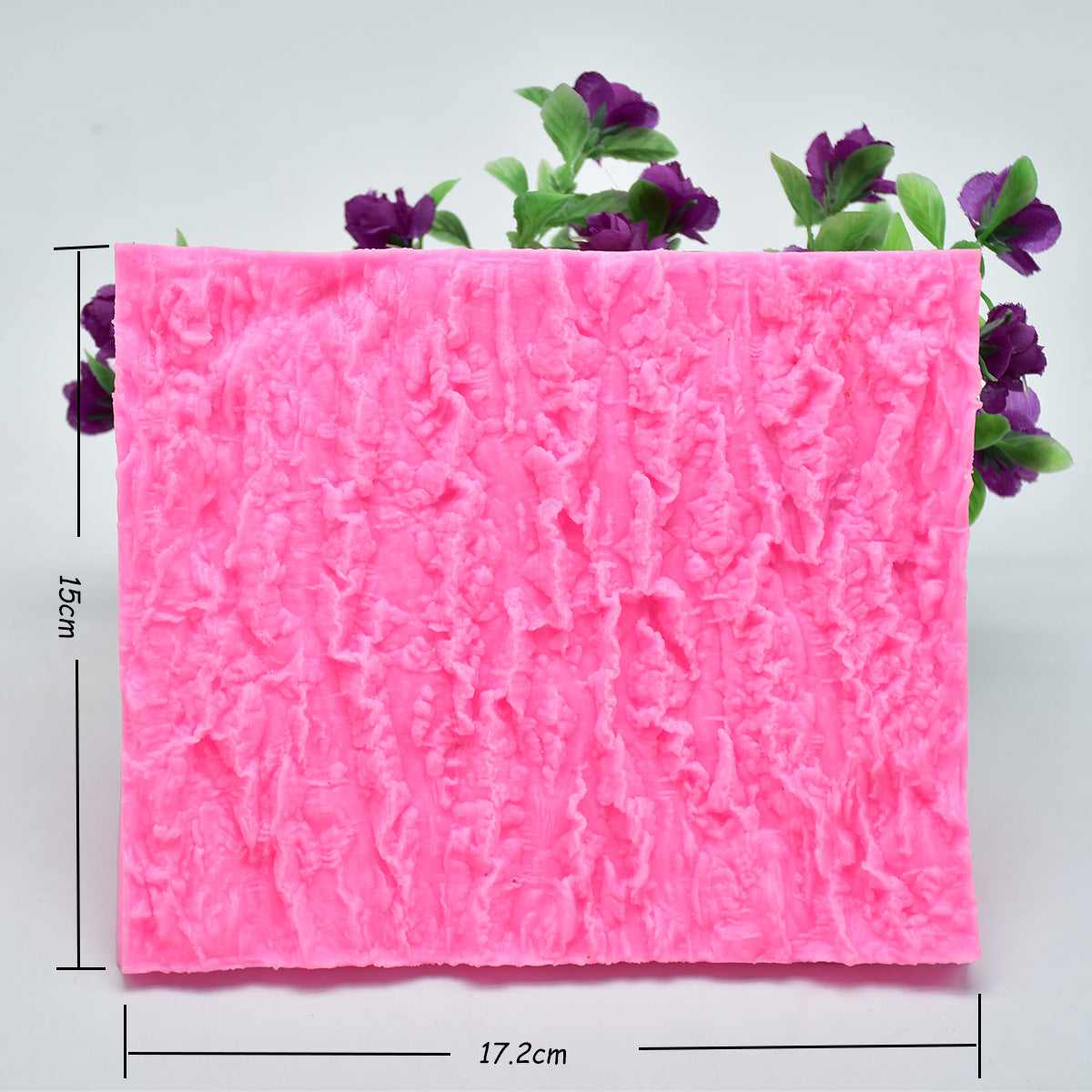 The image shows a pink silicone mat designed for creating a tree bark texture. The mat features a detailed, wrinkled surface that mimics the appearance of tree bark. It measures approximately 15 cm in height and 17.2 cm in width. In the background, there are small purple flowers and green leaves adding some contrast to the composition.