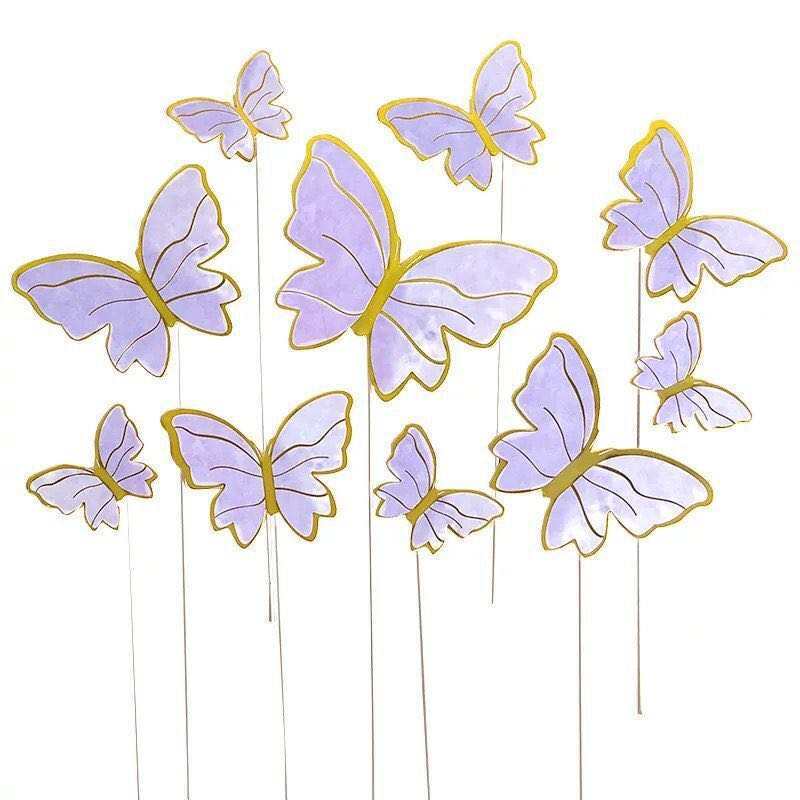 A collection of butterfly cake toppers in various sizes, featuring a soft lavender color with gold outlines. The butterflies are attached to thin white sticks, giving the appearance of them fluttering above a cake. There are a total of five butterflies in different orientations, some larger and some smaller, creating a whimsical and decorative effect.