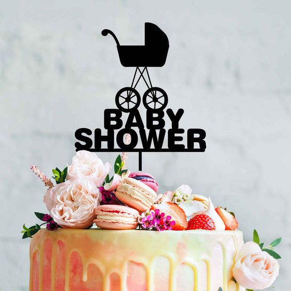 A colorful cake decorated with fresh flowers, macarons, and strawberries. Atop the cake is a black acrylic topper featuring a baby pram design and the words "BABY SHOWER" in bold letters. The background is a light, softly textured surface.