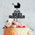 A colorful cake decorated with fresh flowers, macarons, and strawberries. Atop the cake is a black acrylic topper featuring a baby pram design and the words "BABY SHOWER" in bold letters. The background is a light, softly textured surface.