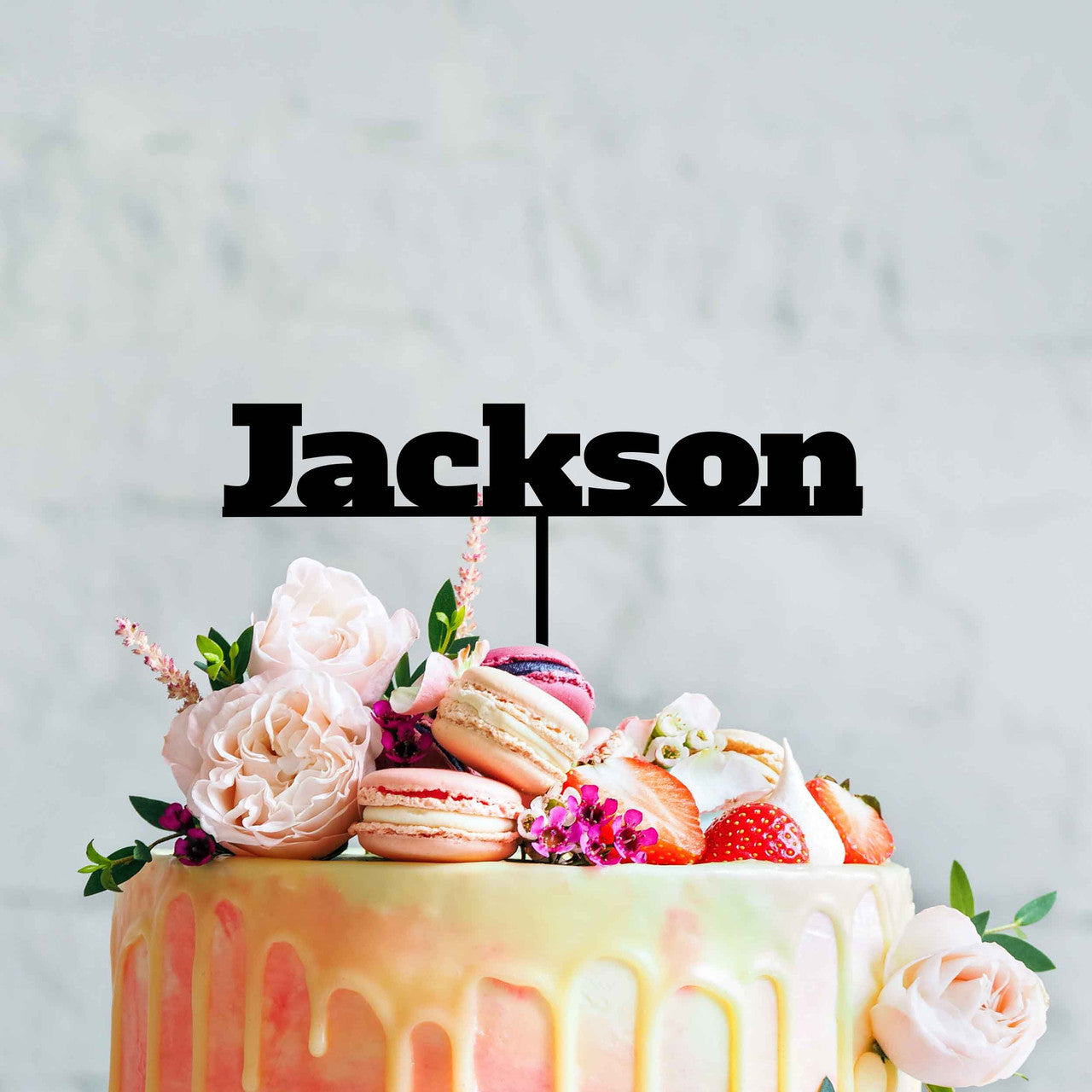A colorful cake topped with an elegant assortment of flowers, macarons, and strawberries. A custom black acrylic cake topper with the name "Jackson" is prominently displayed on top. The cake has a smooth icing with a gradient effect, and the background is a soft, neutral tone.