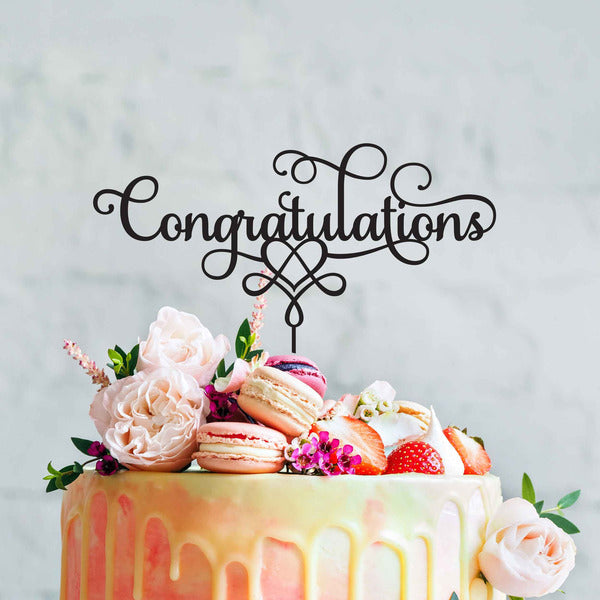 A decorated cake featuring a black acrylic topper that reads "Congratulations" in elegant cursive. The cake is adorned with macarons, strawberries, and various fresh flowers, creating a festive and celebratory appearance. The background is light and neutral, emphasizing the colorful cake and decorations.