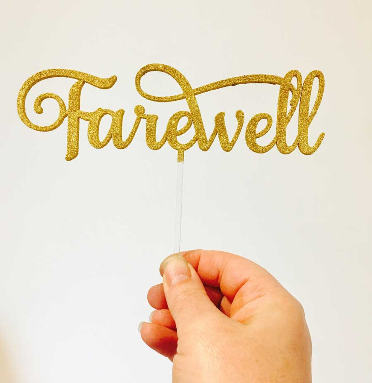 A person holds a gold glittery cake topper that spells "Farewell" in an elegant script. The topper is attached to a clear stick and is set against a plain white background.
