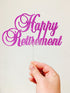 A hand holding a glittery pink acrylic cake topper that reads "Happy Retirement." The topper features decorative cursive lettering and is attached to a clear stem. The background is a plain, light color.