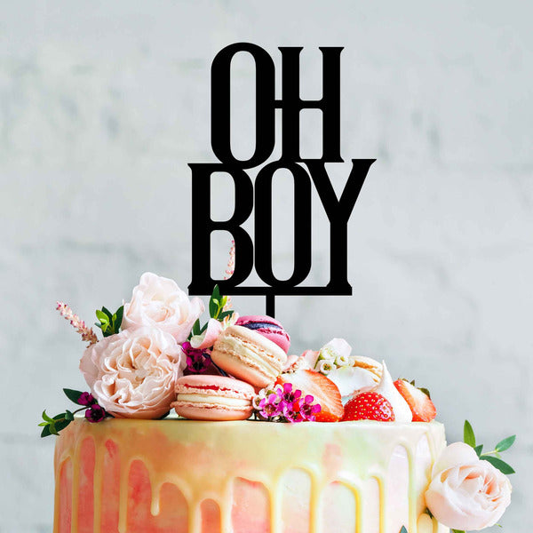 A colorful cake adorned with macarons, strawberries, and flowers, topped with a black acrylic cake topper that reads "OH BOY". The cake has a smooth icing with a dripping effect, and the background is softly blurred.