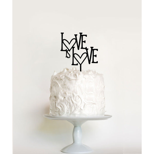 A round white wedding cake with a textured frosting sits atop a simple, white pedestal cake stand. Atop the cake is a black acrylic cake topper that reads "LOVE is LOVE," with decorative hearts incorporated into the lettering. The background is a soft, neutral color, emphasizing the cake and topper.