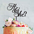 An elegant acrylic cake topper saying "He or She?" in stylish, swirling lettering is prominently displayed on top of a colorful drip cake. The cake features a pale yellow-orange icing with decorative flowers, strawberries, and macarons in shades of pink and purple surrounding the base. The background is softly blurred, focusing on the cake and topper.