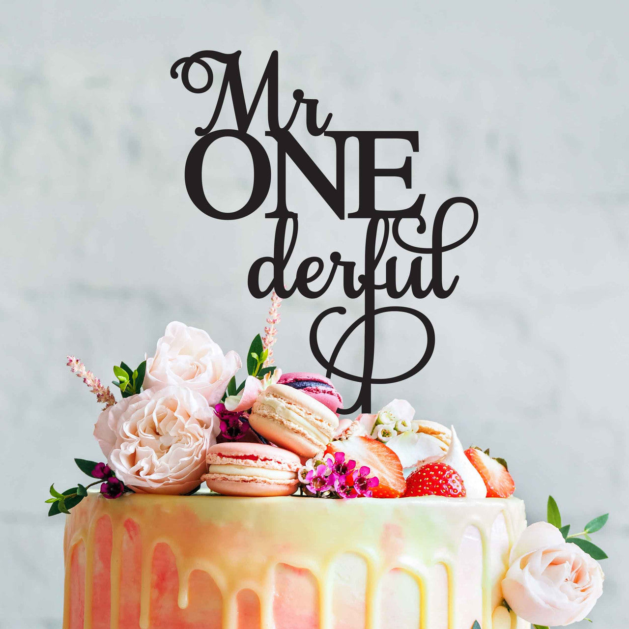 A beautifully decorated cake features an acrylic topper that reads "Mr ONederful" in elegant black lettering. The cake is adorned with pastel-colored icing drips and topped with an arrangement of fresh flowers, macarons, strawberries, and decorative elements. The background is a light, soft color.