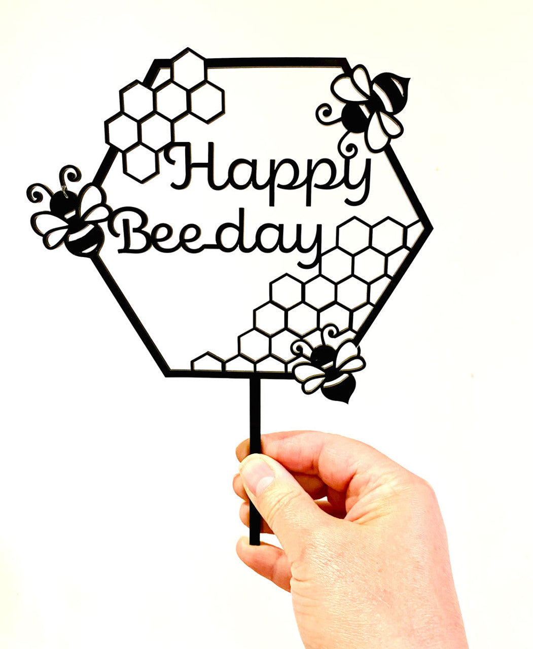 An image of a hand holding a black acrylic cake topper in the shape of a honeycomb. The topper features the text "Happy Beeday" in a playful font, surrounded by decorative bees and hexagonal patterns. The design is whimsical and suited for a bee-themed birthday celebration. The background is plain white, emphasizing the cake topper.