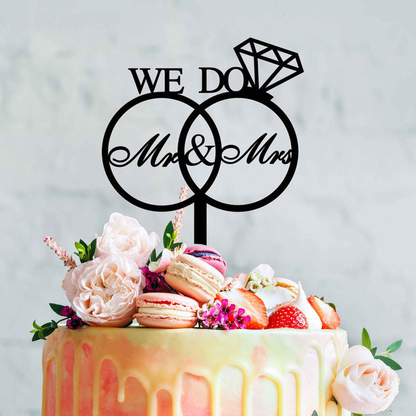 The image features a decorative cake topper designed for weddings. It displays the words "WE DO" above two interlocking circles, each containing "Mr" and "Mrs" in an elegant script font. The topper is adorned with a diamond shape on top. Below the topper, the cake is beautifully decorated with a drip effect and topped with various colorful flowers, macarons, and strawberries, creating an appealing and festive appearance. The background has a light, soft texture that complements the cake.