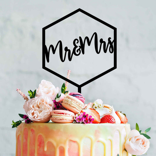 A decorated cake topped with a black acrylic cake topper shaped like a hexagon, featuring the words "Mr & Mrs" in elegant script. The cake has a colorful drip effect and is adorned with fresh flowers, macarons, and strawberries. The background is softly blurred to emphasize the cake and topper.