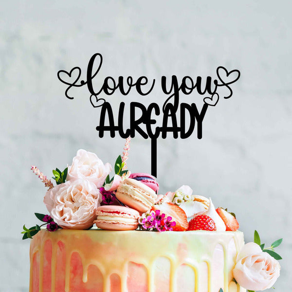 A decorated cake featuring an acrylic topper that reads "love you already" in black cursive letters, adorned with hearts on either side. The cake is pastel-colored with a drip effect and is topped with macarons, strawberries, and various flowers, including light pink roses, against a soft, neutral background.