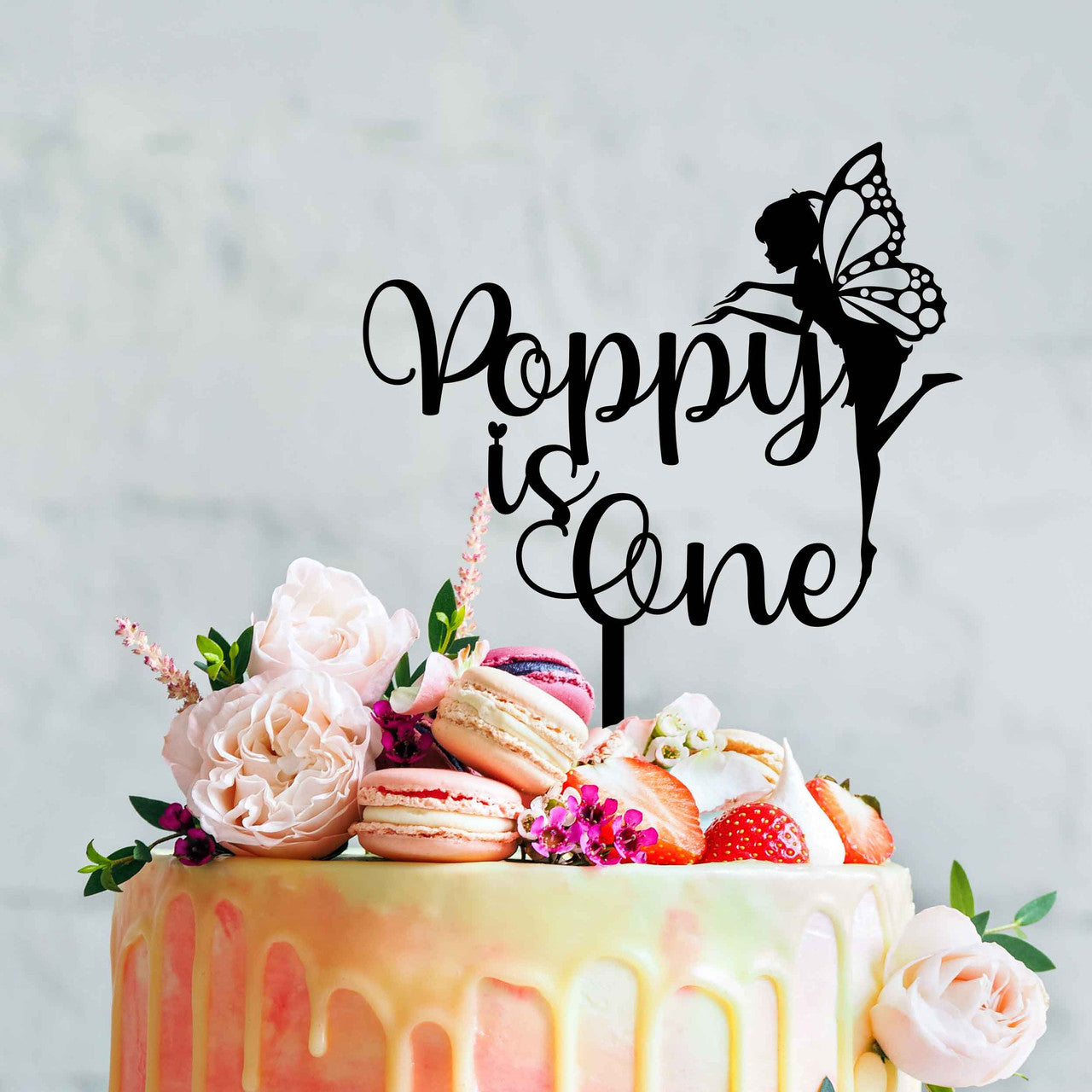 A beautifully decorated cake topped with a black acrylic personalized cake topper featuring the text "Poppy is One" and a silhouette of a fairy. The cake is adorned with pink roses, strawberries, and macarons, set against a soft, light-colored background.