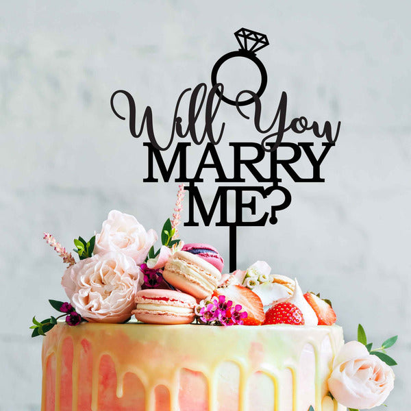 The image features a beautifully decorated cake adorned with a colorful drip and topped with various flowers and macarons. Atop the cake is a black acrylic cake topper that reads "Will You Marry Me?" in an elegant font, accompanied by a diamond ring design. The setting has a soft, neutral background that highlights the cake&