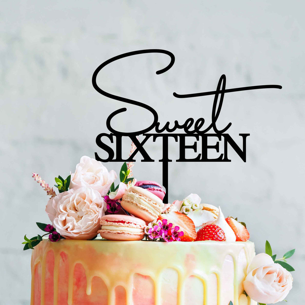 A colorful birthday cake decorated with a glossy drip effect at the sides. Atop the cake is a black acrylic topper that reads "Sweet Sixteen" in a stylish font. The cake is adorned with fresh flowers, including light pink roses, along with vibrant strawberries and colorful macarons, adding a festive touch. The background is a soft, neutral tone, enhancing the cake&