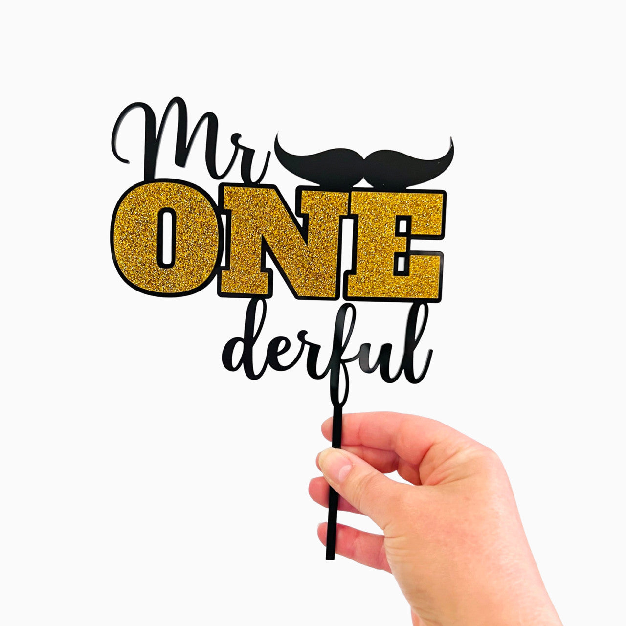 A hand is holding a cake topper that reads "Mr ONE derful." The word "Mr" is in a stylish font, while "ONE" is prominently displayed in bright gold glitter. Below "ONE," the word "derful" is in a cursive, black font. A small black mustache is positioned above the word "ONE." The overall design is festive and suitable for a birthday celebration, particularly for a first birthday. The backdrop is plain white.