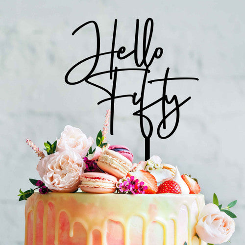 A colorful cake decorated with flowers, macarons, and strawberries, topped with a black acrylic cake topper that reads ?Hello Fifty? in a modern script font. The background is a light, neutral color that highlights the cake&