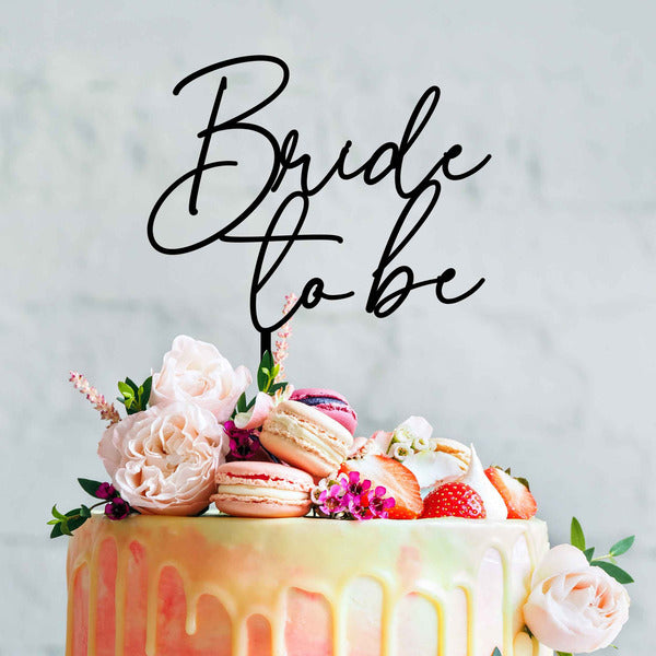 The image features a beautifully decorated cake adorned with various toppings. At the top, there is a stylish acrylic cake topper that reads "Bride to be" in elegant, cursive lettering. Surrounding the topper are pink and white flowers, colorful macarons, and fresh strawberries, adding a festive touch to the cake. The cake itself has a gradient frosting with a smooth, drippy design, set against a light, neutral background.