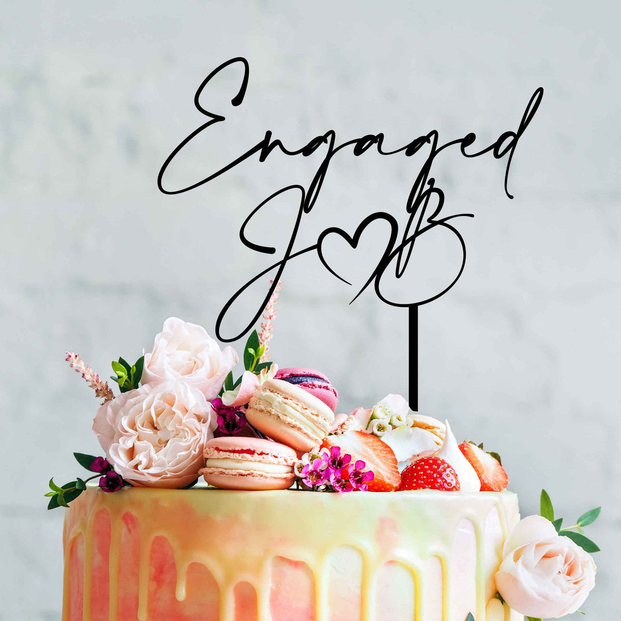 A decorative wedding cake featuring a smooth, pastel-colored icing with a drip effect at the edges. Topped with fresh flowers, strawberries, and macarons. Atop the cake is a black acrylic topper that reads "Engaged" with a heart symbol and the initials "J" and "B". The background is a soft, neutral tone.