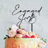 A decorative wedding cake featuring a smooth, pastel-colored icing with a drip effect at the edges. Topped with fresh flowers, strawberries, and macarons. Atop the cake is a black acrylic topper that reads "Engaged" with a heart symbol and the initials "J" and "B". The background is a soft, neutral tone.
