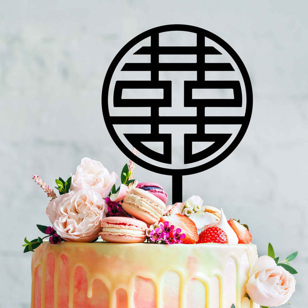 A colorful cake topped with a black acrylic cake topper featuring the "double happiness" Chinese symbol. The cake is decorated with pink macarons, strawberries, and fresh flowers, including pale pink roses and green leaves. The background is softly blurred, highlighting the cake and its decorations.