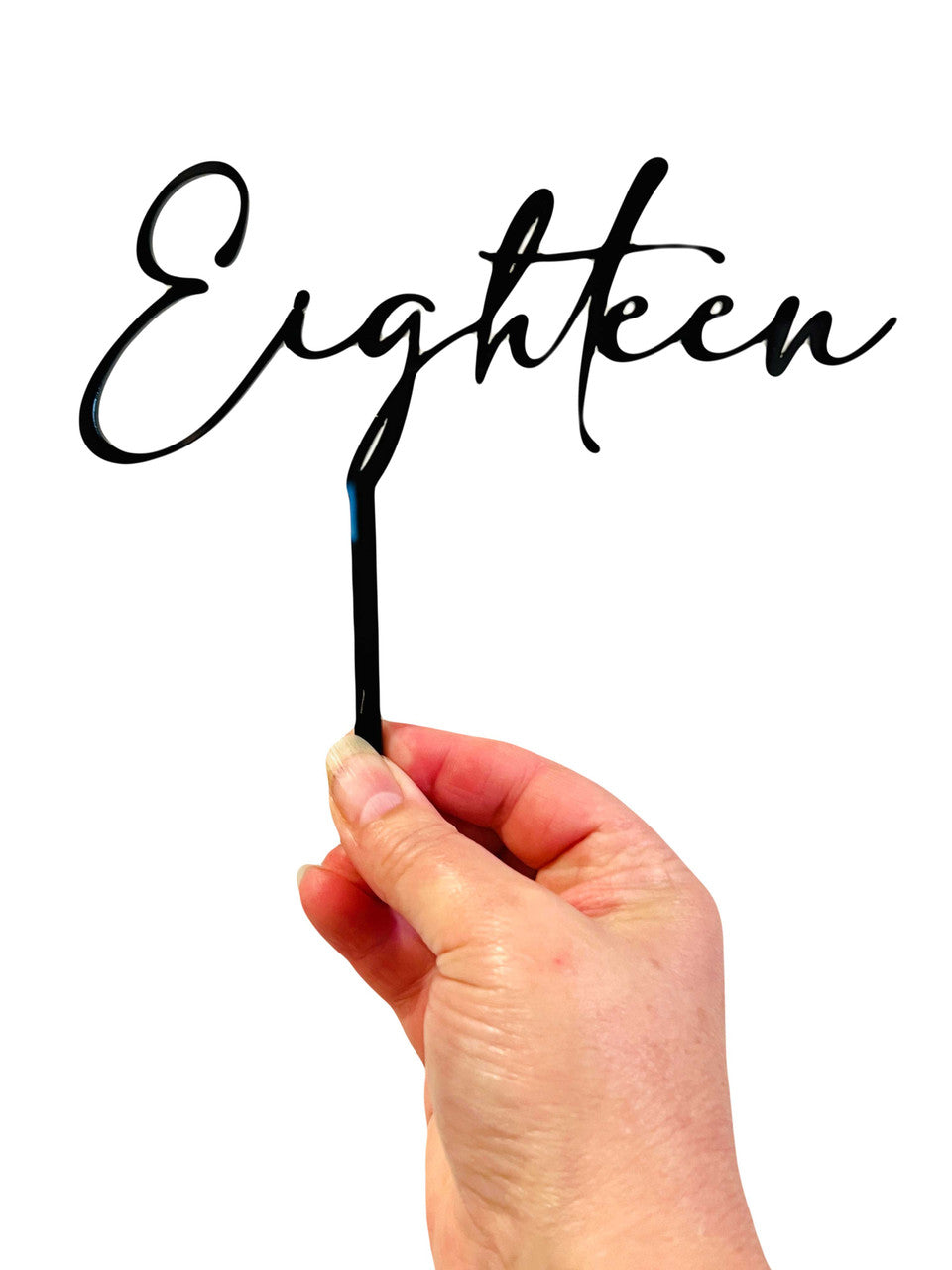 A person is holding a black acrylic cake topper that features the scripted word "Eighteen." The topper has a thin stick for insertion into a cake and is displayed against a plain white background. The hand holding the topper has light-colored nails.