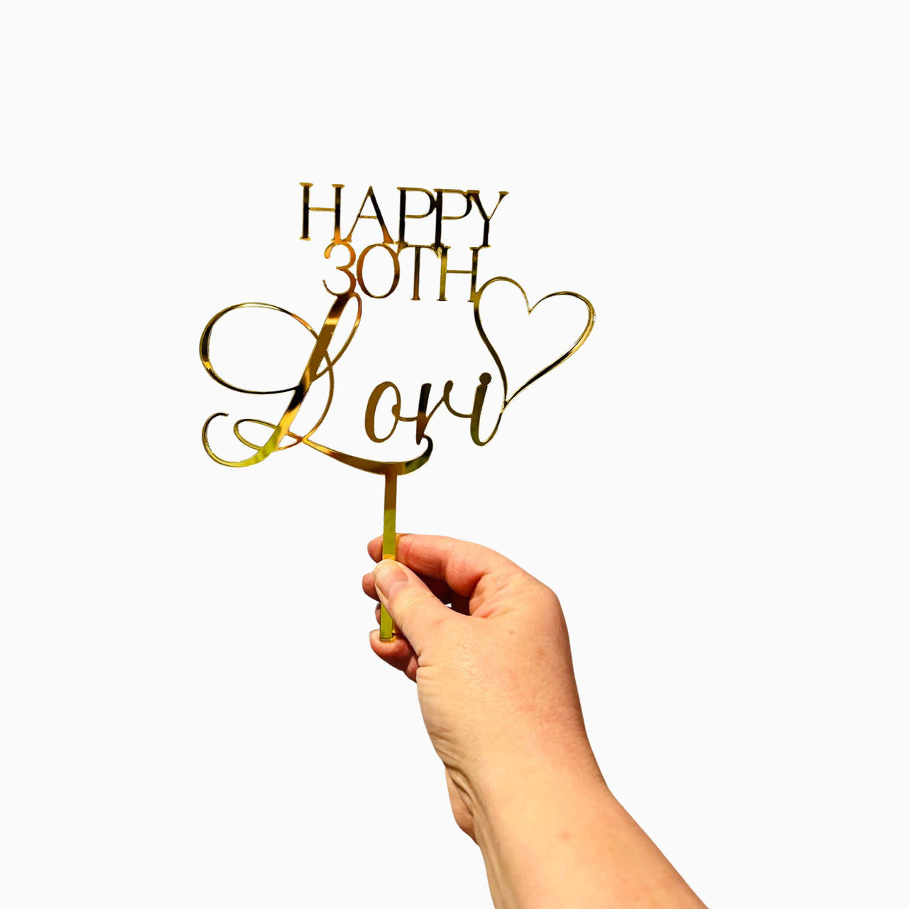 A hand is holding a gold acrylic cake topper with the text "HAPPY 30TH Lori" in an elegant, cursive font. The design includes a decorative heart next to the name. The background is plain white, highlighting the shiny gold finish of the topper.
