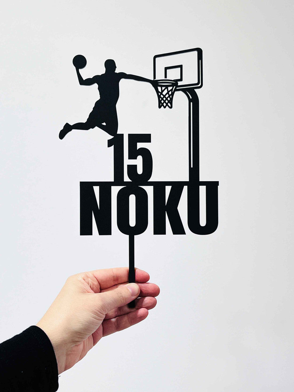 An acrylic cake topper featuring a silhouette of a basketball player making a slam dunk. The design includes the number "15" and the name "NOKU" in bold, black letters. The topper is being held by a hand, with a plain white background.