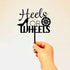 A hand is holding a black acrylic cake topper that features the words "Heels or WHEELS." The design includes a stylized high heel shoe next to the word "Heels" and a wheel icon next to the word "WHEELS." The background is plain and neutral in color, allowing the topper to stand out prominently.