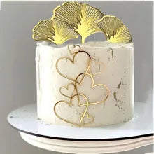 A round white cake topped with a decorative acrylic topper featuring gold ginkgo leaves. The cake showcases smooth white icing and is adorned with a design of intertwined gold hearts on the front. The cake is placed on a white cake stand.