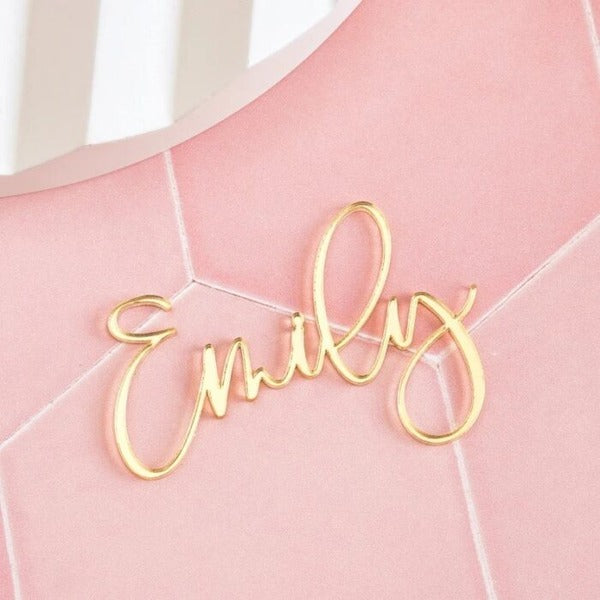 The image features a personalized acrylic cake topper with the name "Emily" written in an elegant, cursive gold font. The topper is placed on a soft pink background, which enhances its decorative appeal. The design emphasizes a stylish and customized look suitable for a cake decoration.